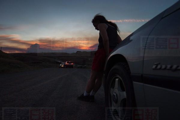 A woman who was evacuated from Cache Creek rests on the front of her car along the Trans-Canada Highway in Savona, B.C., as smoke from a wildfire burning near Ashcroft rises in the distance at sunset on Friday July 7, 2017. THE CANADIAN PRESS/Darryl Dyck