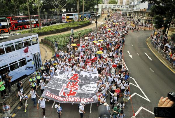 People stand on a pavement watch protesters holding banner and placards march on a street in a rally against the proposed amendments to extradition law in Hong Kong Sunday, June 9, 2019. The extradition law has aroused concerns that this legislation would undermine the city's independent judicial system as it allows Hong Kong to hand over fugitives to the jurisdictions that the city doesn't currently have an extradition agreement with, including mainland China, where a fair trial might not be guaranteed. (AP Photo/Kin Cheung)