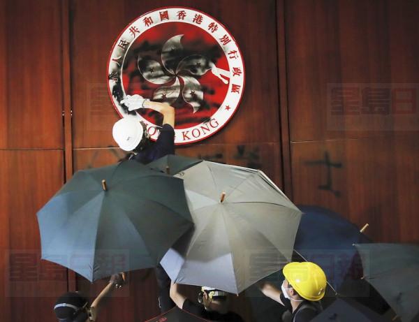 Protesters with umbrellas help a protester defaces the Hong Kong emblem after they broke into the Legislative Council building in Hong Kong, Monday, July 1, 2019. Protesters in Hong Kong took over the legislature's main building Monday night, tearing down portraits of legislative leaders and spray painting pro-democracy slogans on the walls of the main chamber.(AP Photo/Kin Cheung)