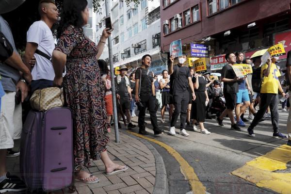 Tourists watch as protesters march in an area popular with Chinese tourists for its pharmacies and cosmetic shops, in Hong Kong, Saturday, July 13, 2019. Several thousand people marched in Hong Kong on Saturday against traders from mainland China in what is fast becoming a summer of unrest in the semi-autonomous Chinese territory. (AP Photo/Kin Cheung)