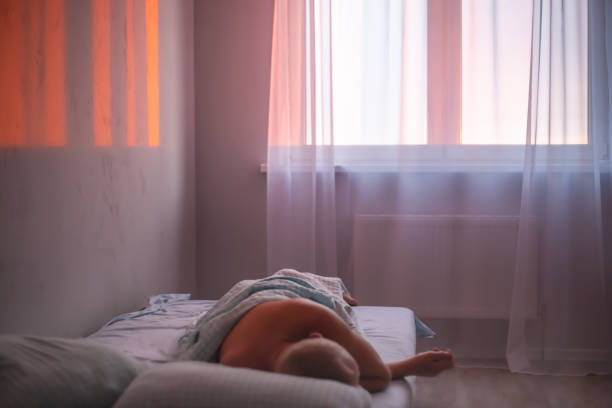 In the early morning, the man sleeps in bed. Sunbeams at dawn fall on the wall in the apartment through a window with transparent tulle. Atmospheric mood.